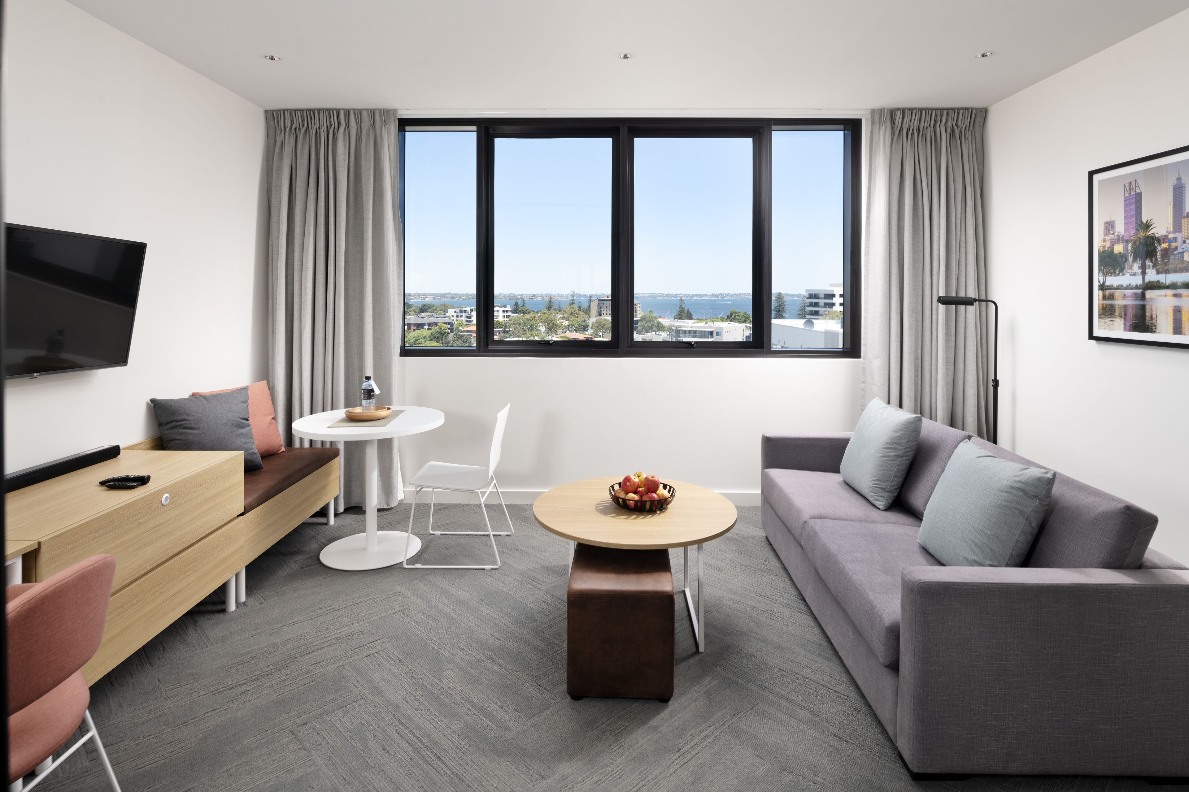 A modern hotel room with a sofa, a dining table, a TV, and a large window with a city view.