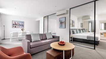 Una habitación de hotel moderna con un sofá gris, una mesa de centro redonda de madera, una silla roja, una cama con ropa de cama gris y un espejo grande.