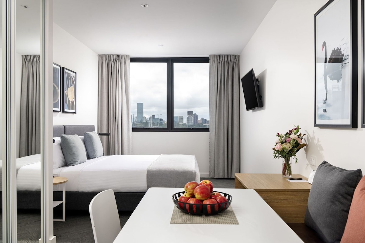 A hotel room with a bed, a desk with a bowl of fruit, a chair, a table, a TV, and a window with curtains.
