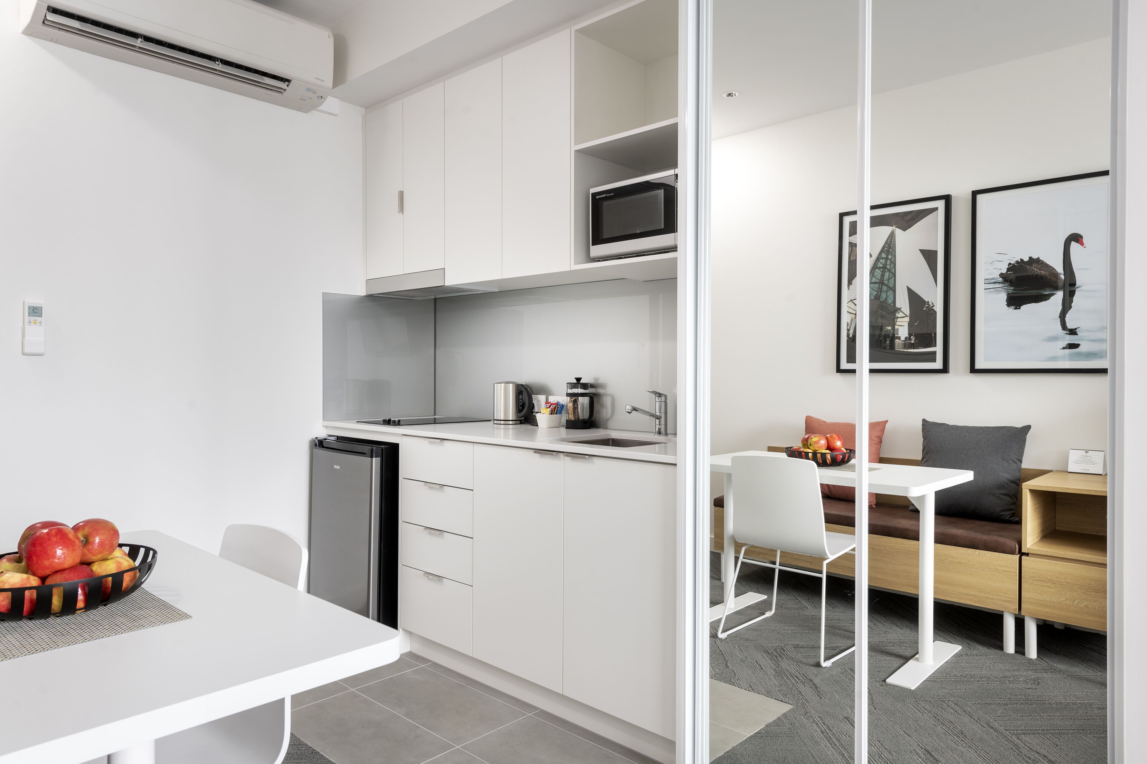 A modern kitchen with white cabinetry, a microwave, and a sink. There is a dining area with a white table and chairs, and a sofa with cushions.