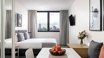 A hotel room with a bed, a desk, a chair, a dining table with fruit, and a city view.