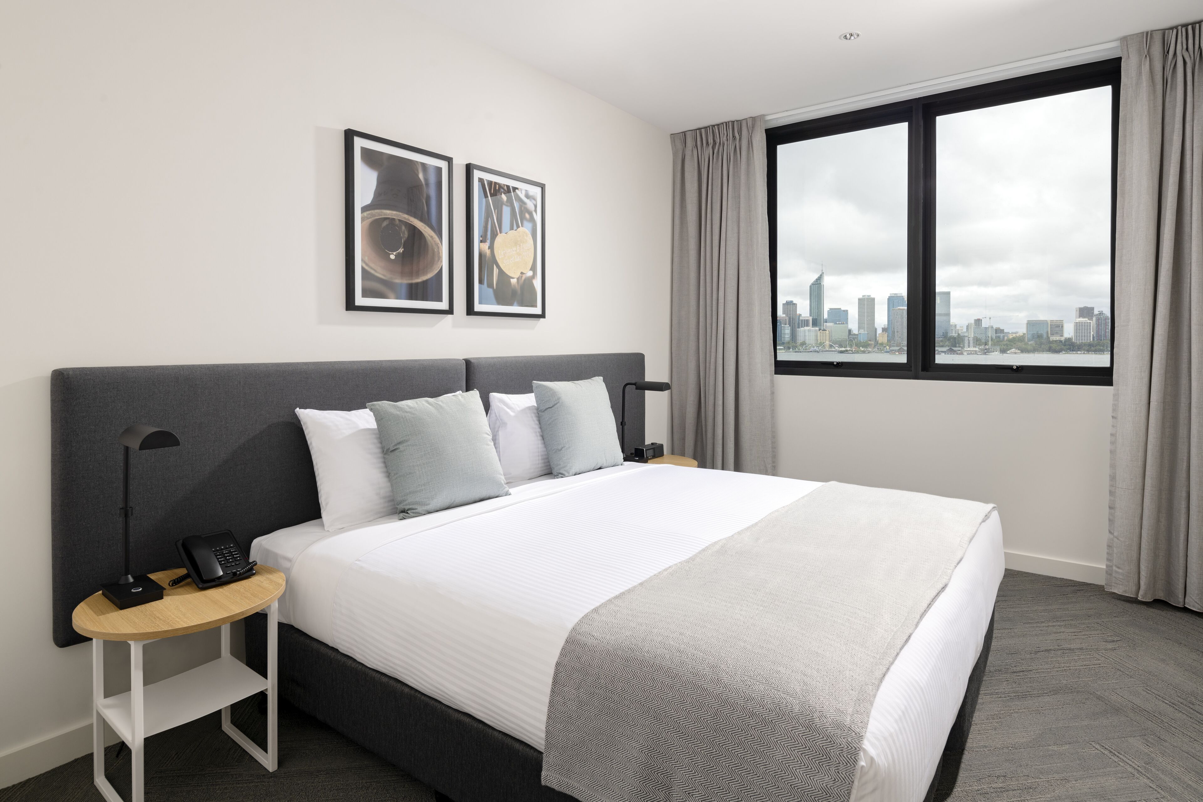 A modern hotel room with a large bed, a bedside table with a phone, and a city view through a large window.