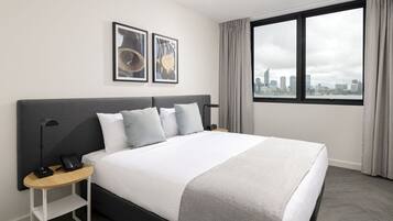 A modern hotel room with a large bed, a bedside table with a phone, and a city view through a large window.