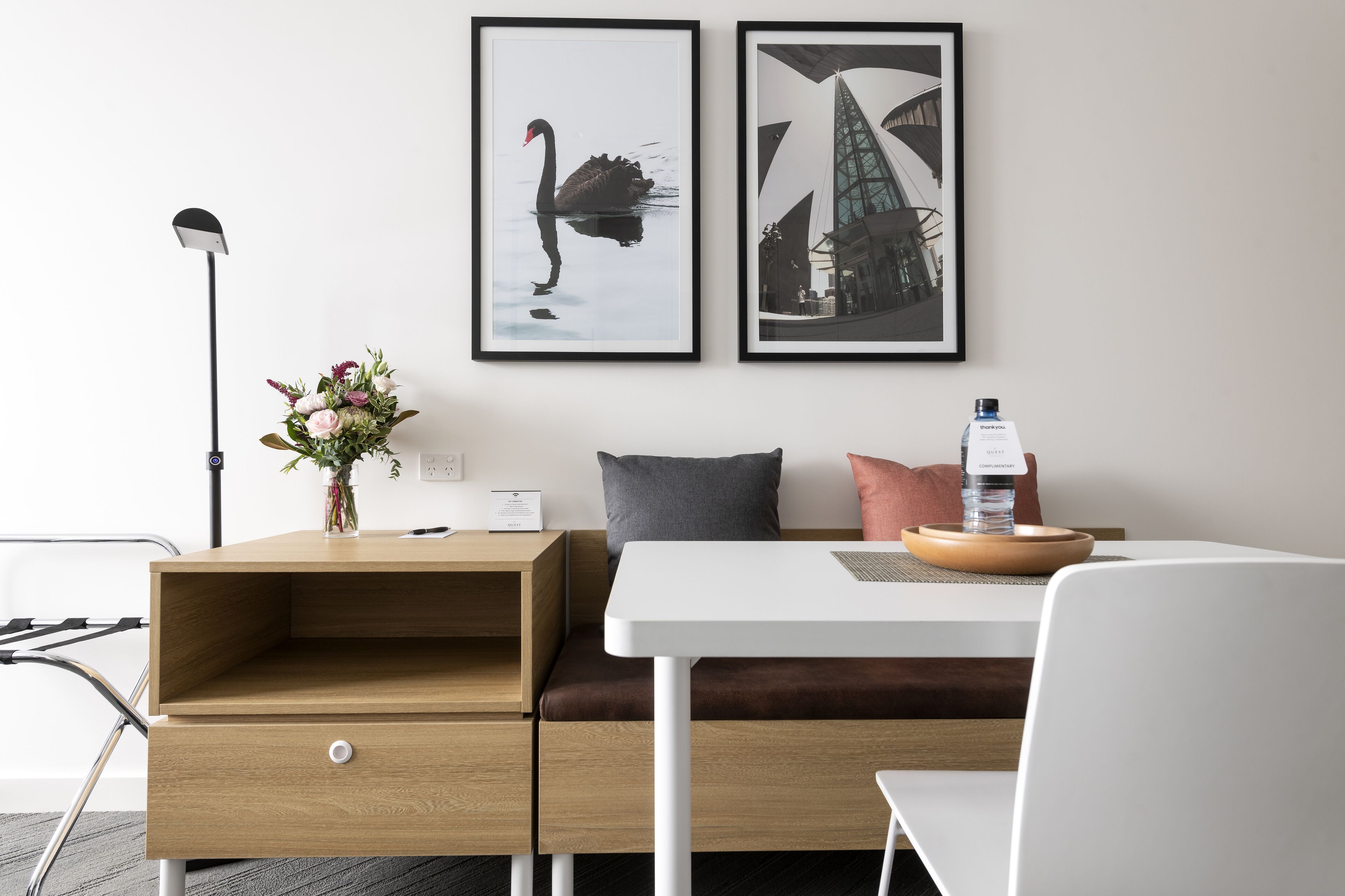 Habitació d'hotel moderna amb escriptori de fusta, una taula blanca i un sofà. Hi ha dos quadres amb marc a la paret, un vas amb flors a l'escriptori i una ampolla d'aigua a la taula.