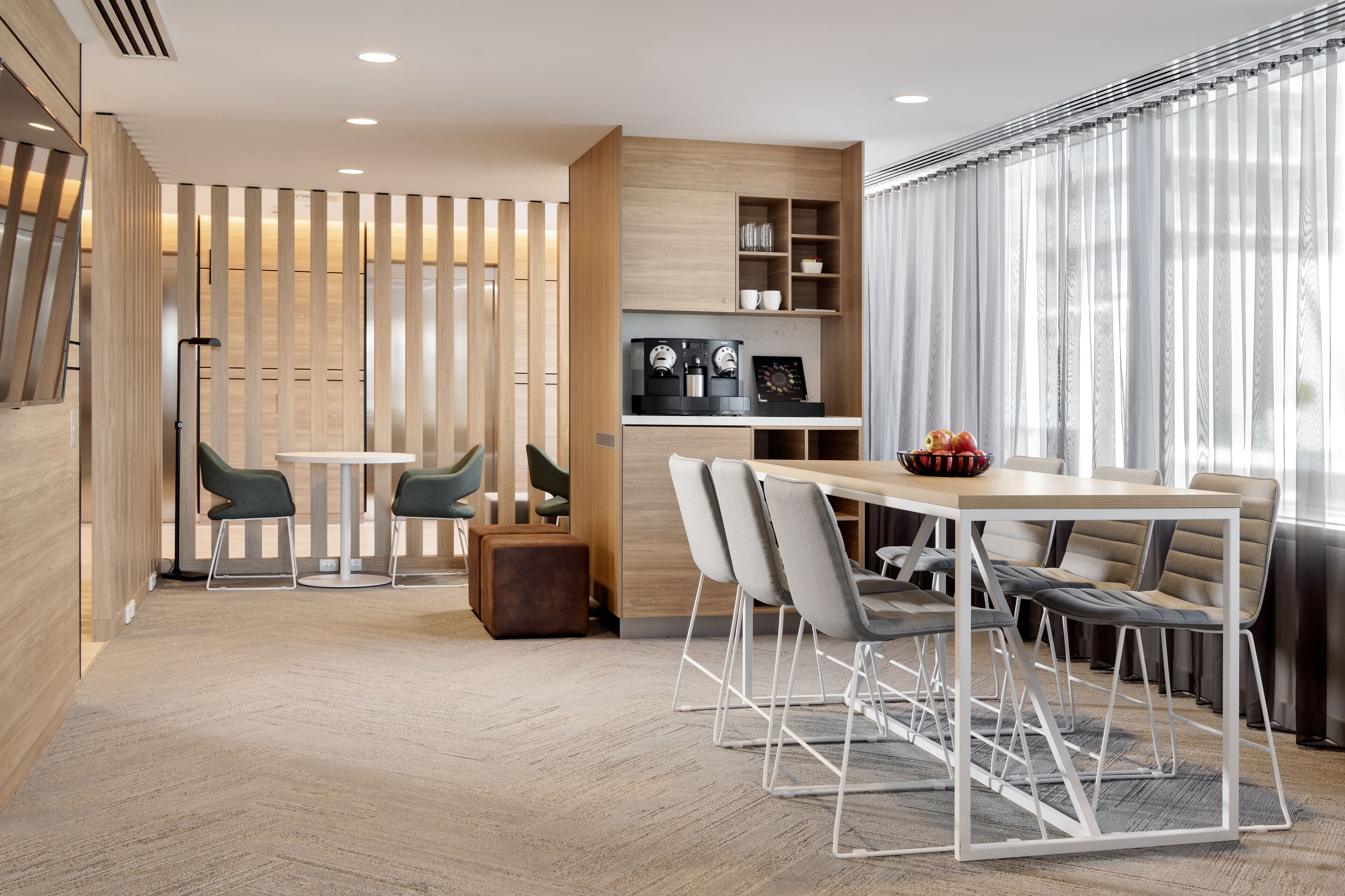 Una habitación de hotel moderna con piso de madera, un área de comedor con mesa y sillas, una kitchenette y un amplio ventanal con cortinas.
