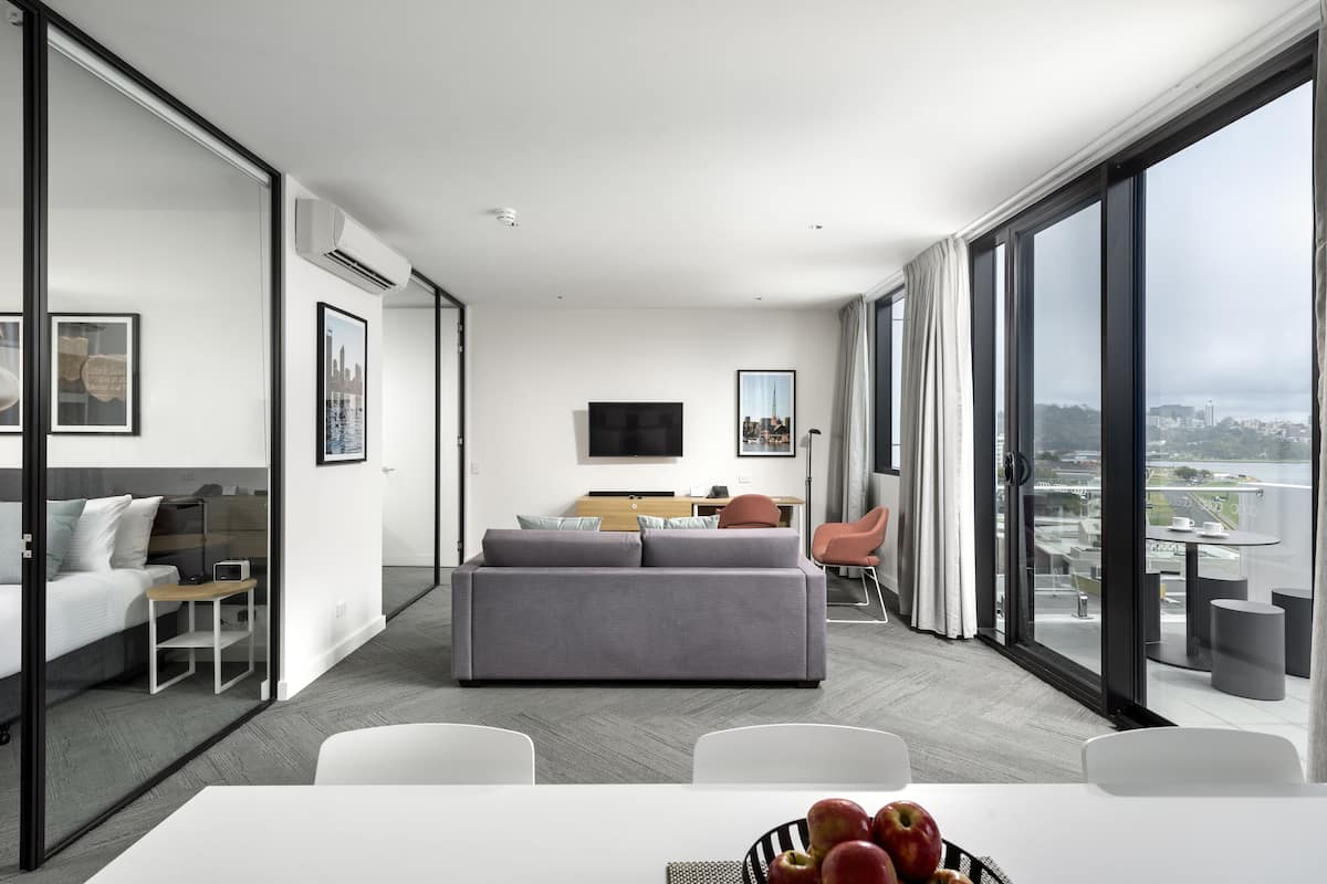 A modern hotel room with a grey sofa, white dining table, and large windows offering a city view.