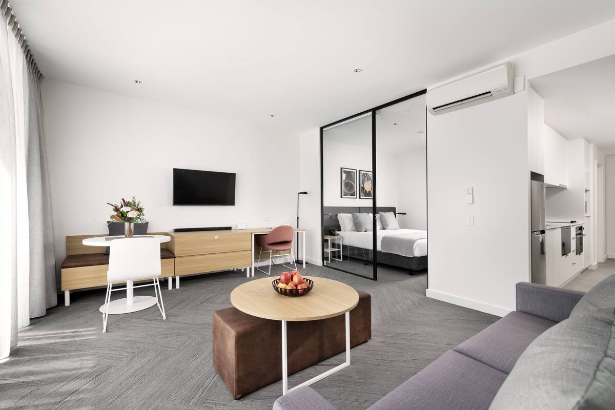 A modern, well-lit room with a sofa, a dining table with a bowl of fruit, a desk with a chair, a bed, and a TV mounted on the wall.
