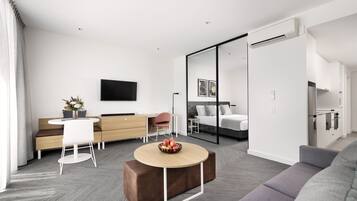 A modern, well-lit room with a sofa, a dining table with a bowl of fruit, a desk with a chair, a bed, and a TV mounted on the wall.