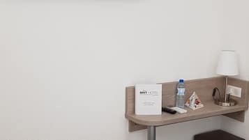 A hotel room with a bed, a TV mounted on the wall, a bedside table with a lamp, a trash bin, and a water bottle.