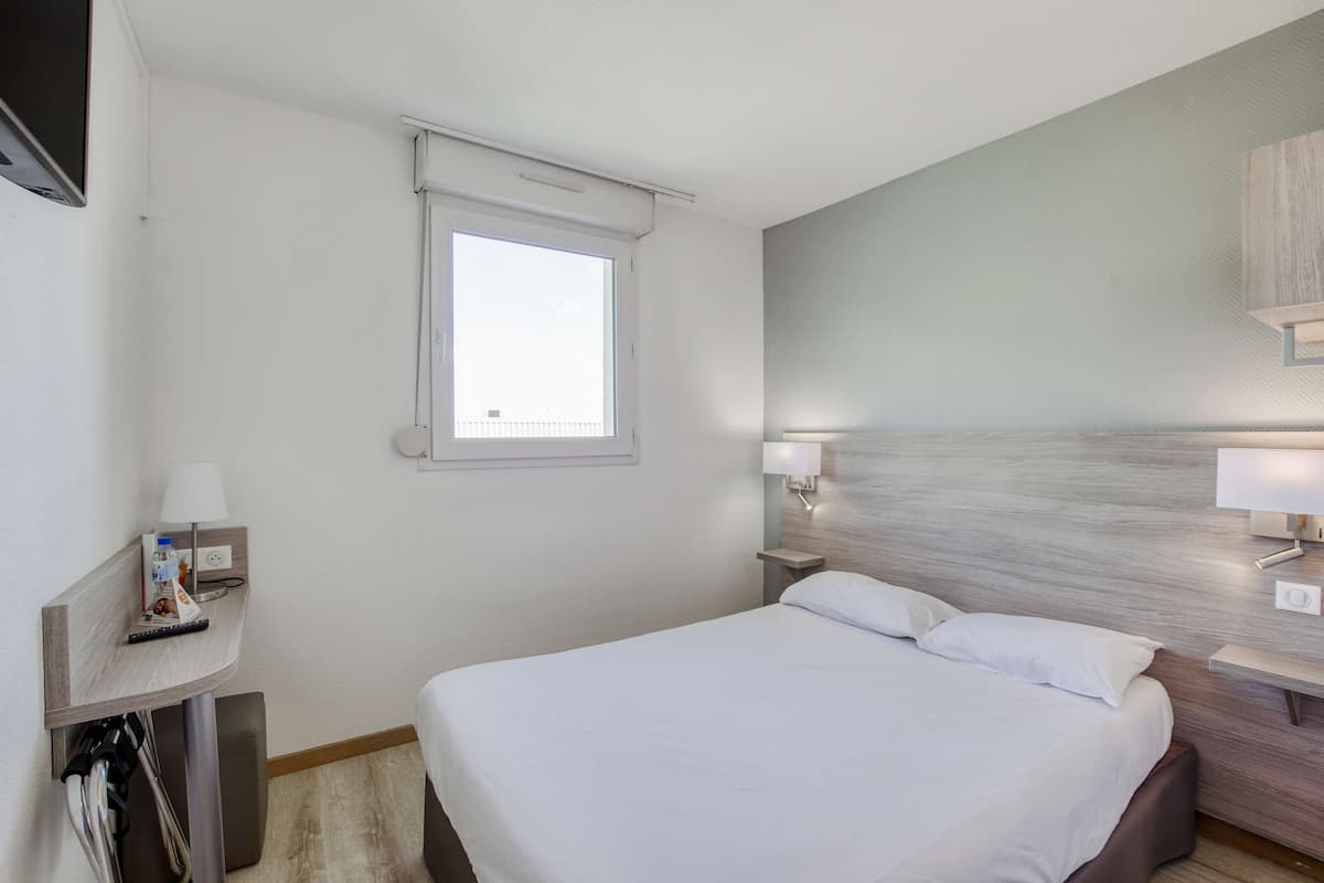 Ein Hotelzimmer mit Bett, Nachttischlampen, einem Fenster und einem kleinen Tisch mit Lampe und Fernbedienung.