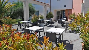 Un espace extérieur aménagé en terrasse, avec plusieurs tables et chaises, entouré de plantes en pot et bordé par un bâtiment doté d’un toit en tuiles.