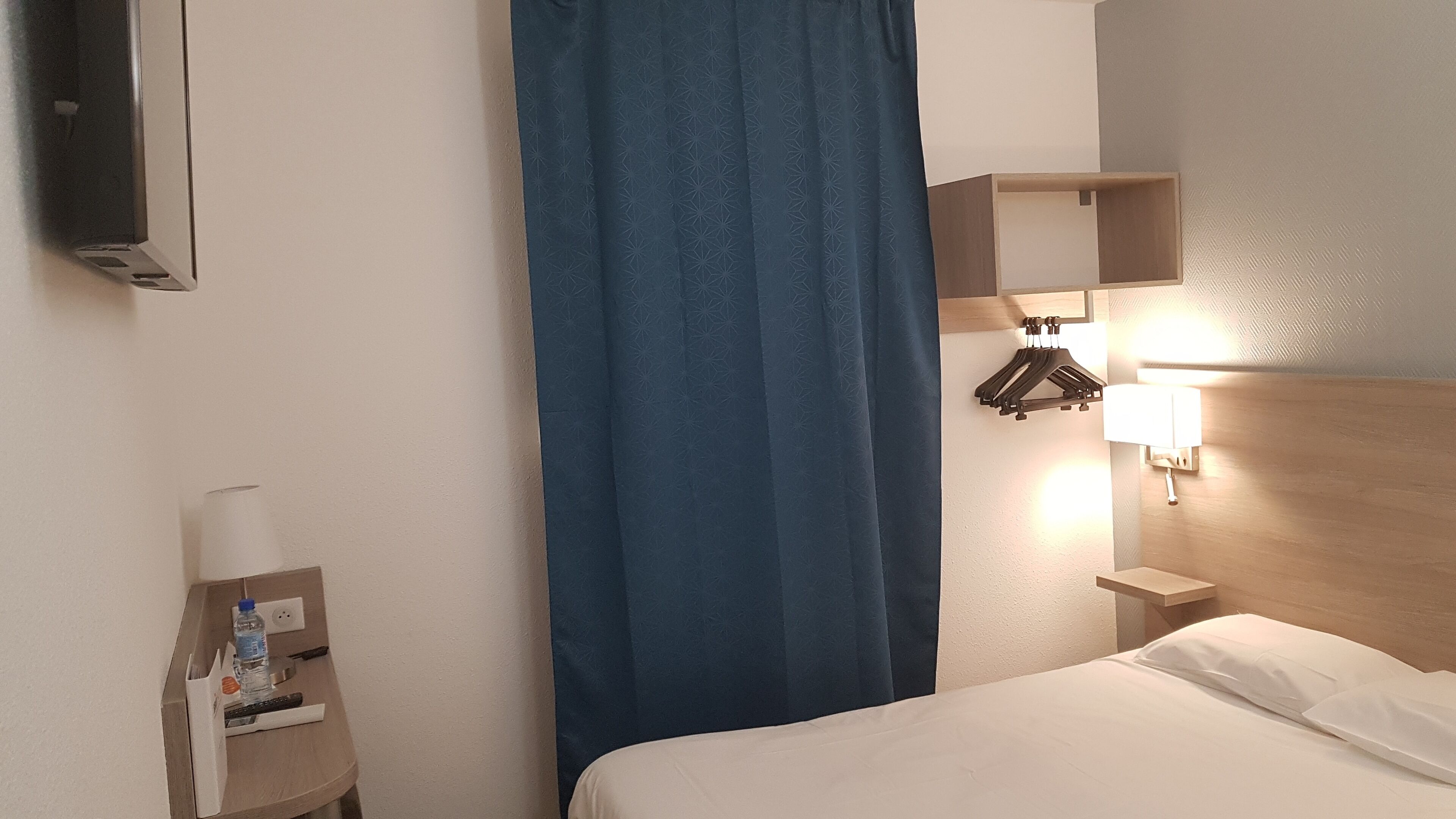 Una camera d'albergo con un letto, un comodino con una lampada, una TV a parete, una tenda blu e un poggiatesta in legno.