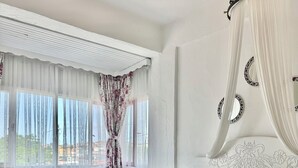 A bedroom with a large bed, a bedside table, a nightstand, a window with curtains, and a chandelier. - Aisem By Hire Alacati (Cesme)