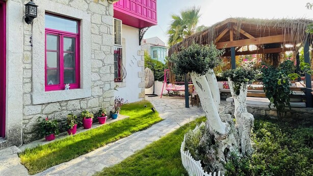 A two-story house with a stone facade and pink accents, a well-maintained lawn, and a white picket fence. - Aisem By Hire Alacati (Cesme)