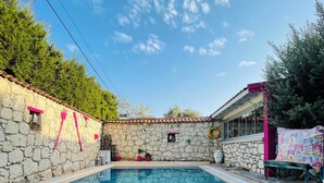 A swimming pool with stone walls and a tiled floor, surrounded by greenery and a building with a tiled roof. - Aisem By Hire Alacati (Cesme)