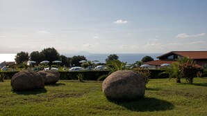 A grassy area with large rocks, a building with a red roof, and umbrellas in the background. - Agriturismo Ninea (Ricadi)