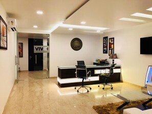 A modern office interior with a reception desk, two office chairs, a mounted TV, and a decorative wall piece. - Vivid Boutique Hotel (Gurugram)