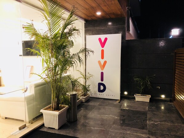 A hotel entrance with a modern design, featuring a reception desk, potted plants, and a prominent sign with the name 'VIVID HOTEL'.