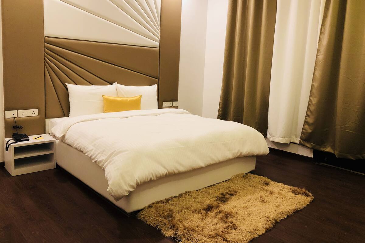 Una cama bien hecha con almohadas blancas y amarillas, un cabecero marrón y una mesita de noche de madera con un teléfono y una pequeña estantería.