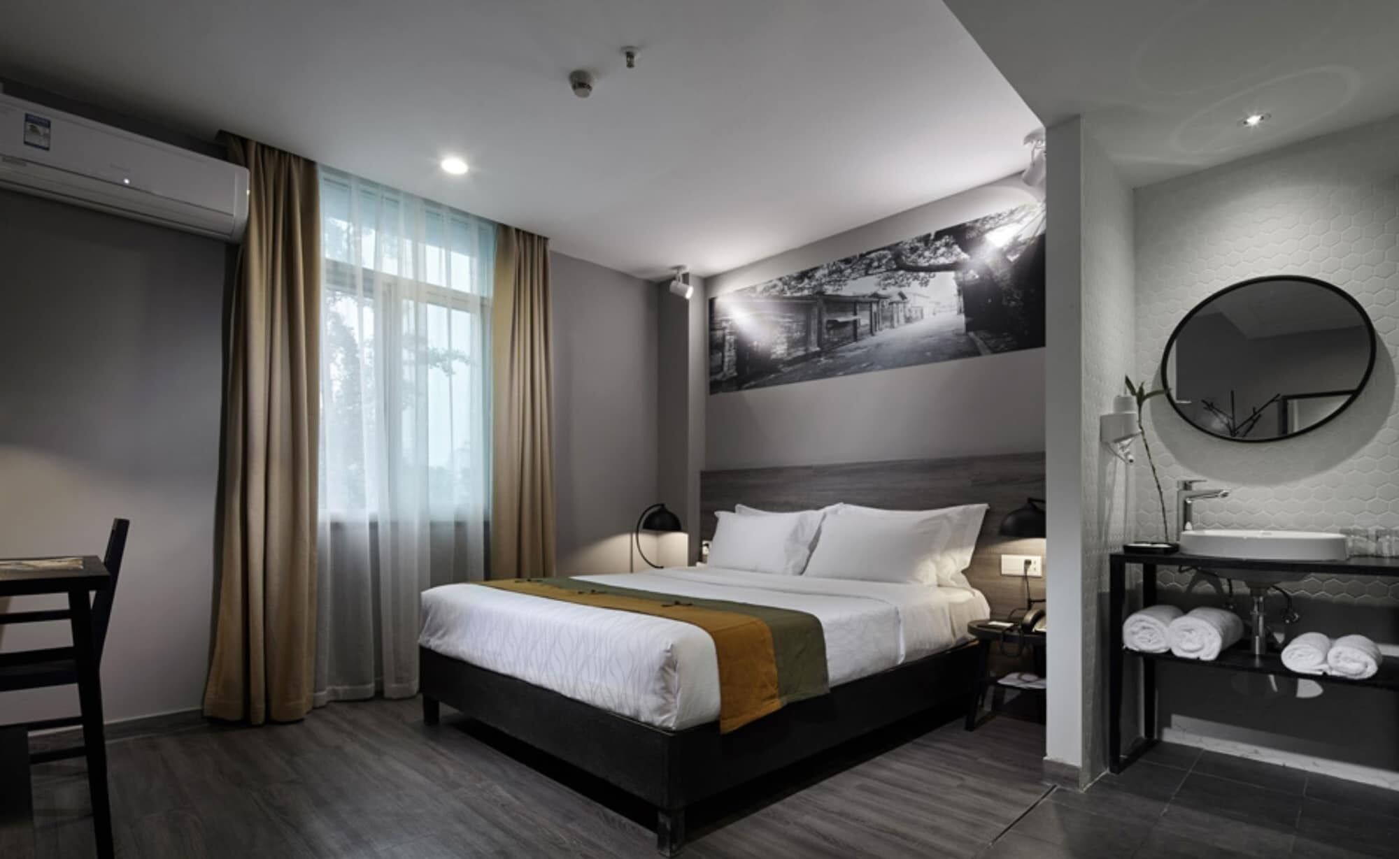 Une chambre d’hôtel moderne dotée d’un grand lit, d’un bureau, d’une chaise, d’une salle de bain avec un grand miroir rond et un lavabo, ainsi qu’une œuvre d’art accrochée au mur.