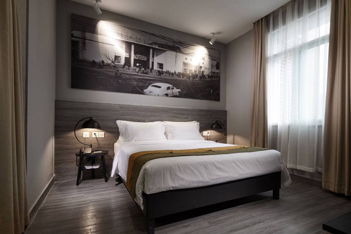 Une chambre d’hôtel moderne avec un grand lit, deux tables de chevet, une œuvre d’art accrochée au mur et une grande fenêtre avec des rideaux.