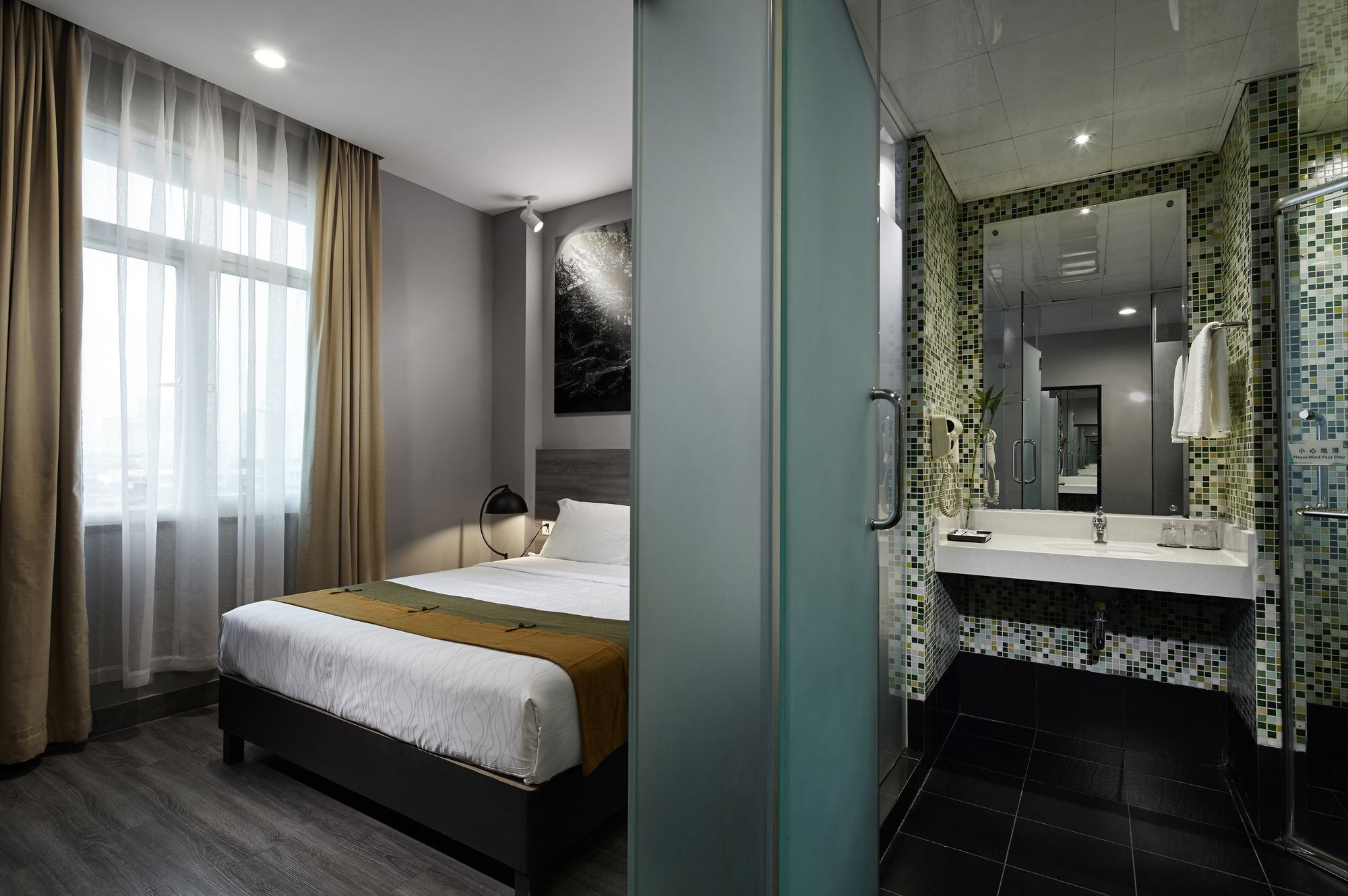 Una habitación de hotel con cama, escritorio, silla, ventana con cortinas y baño con lavabo, espejo y ducha.