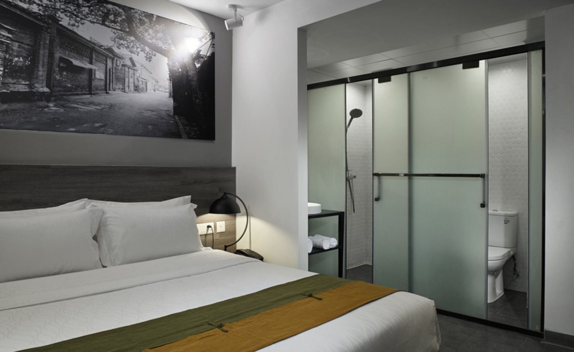 A hotel room with a bed, a nightstand, a lamp, a framed picture on the wall, and a sliding glass door leading to a bathroom.