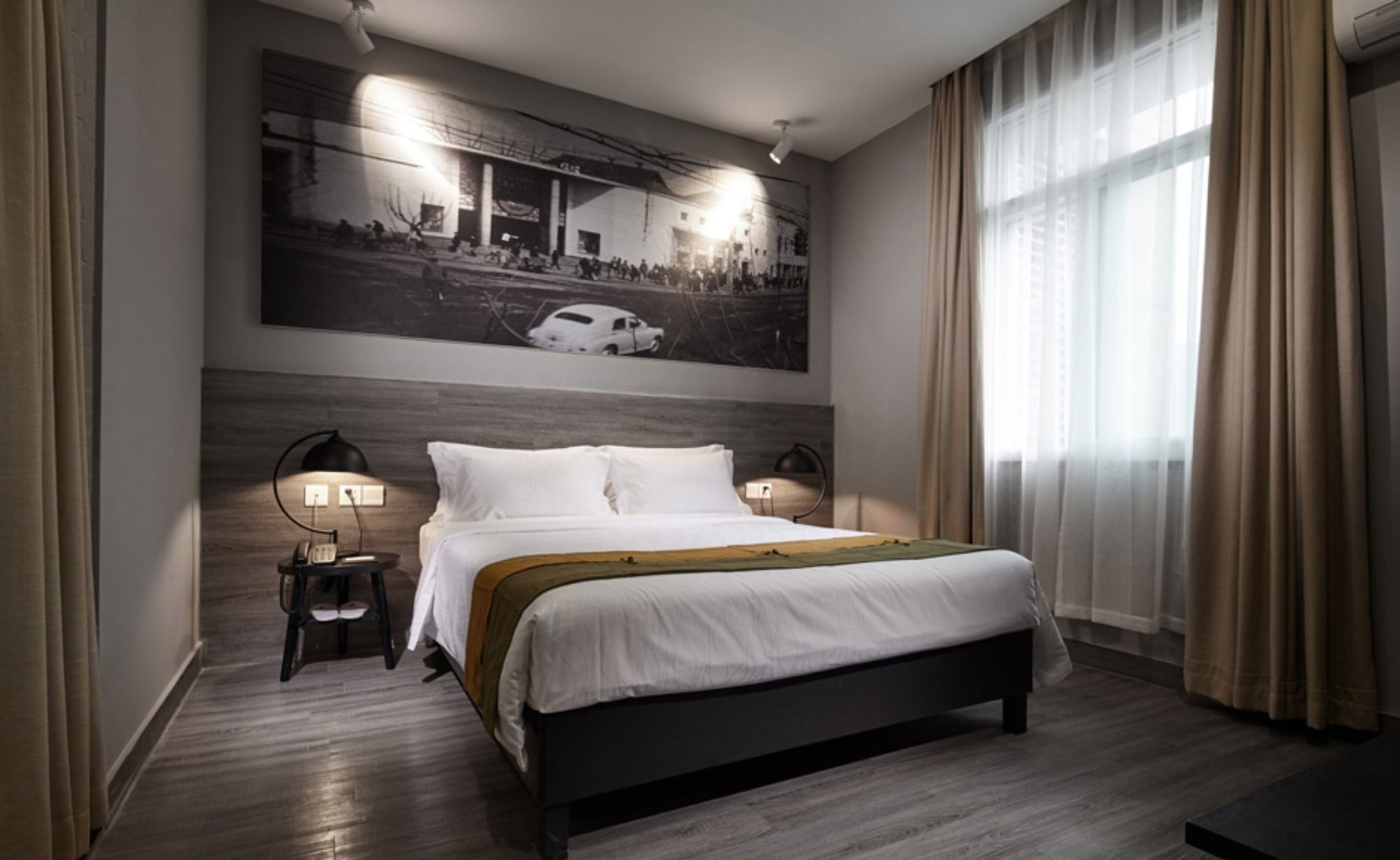 A modern hotel room with a large bed, two bedside tables with lamps, a large window with curtains, and a cityscape painting on the wall.