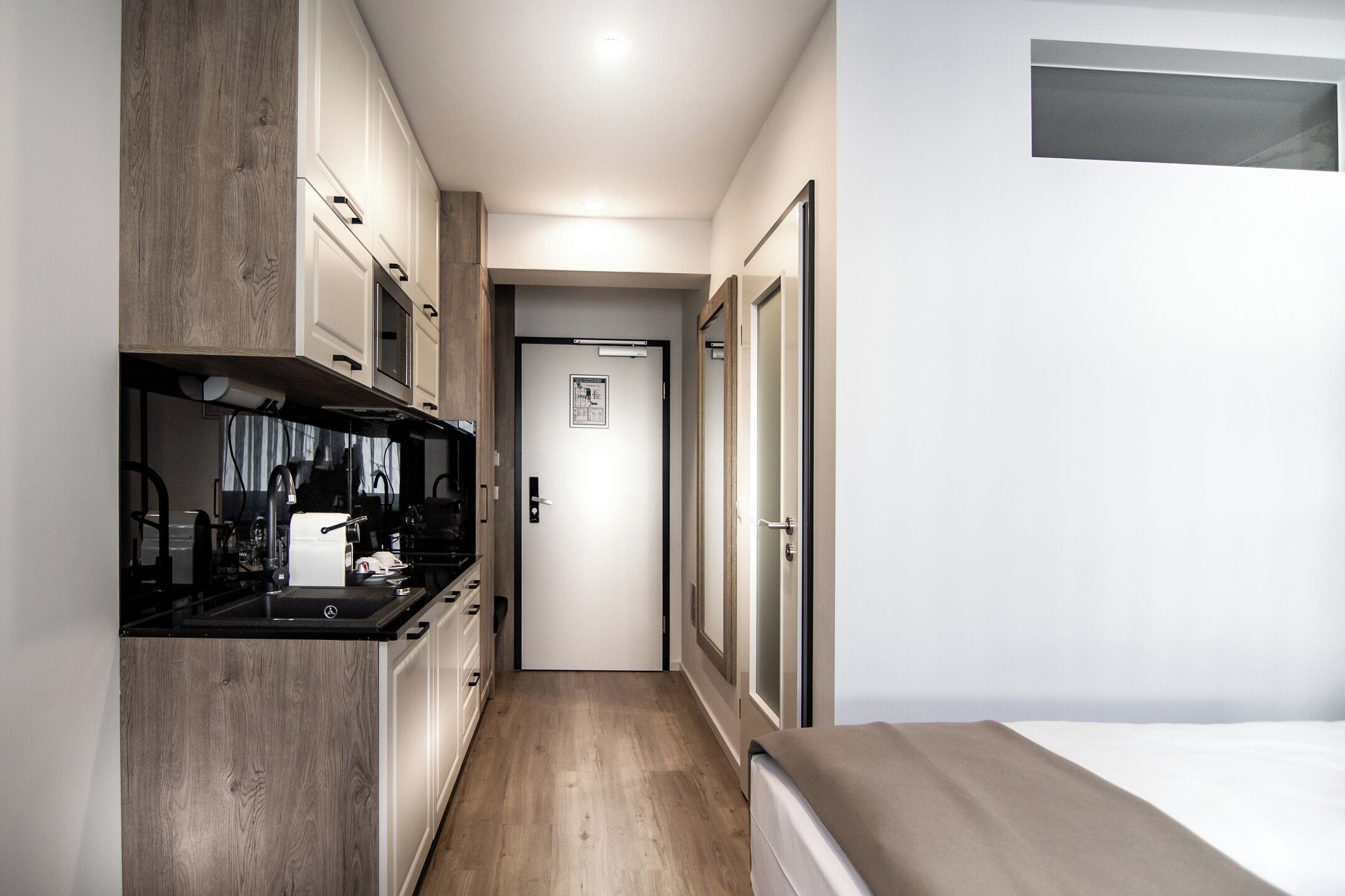 Un couloir étroit, avec une kitchenette sur la gauche, une porte au fond et un lit sur la droite.