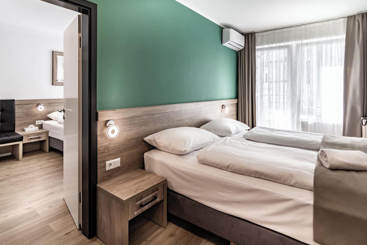Une chambre d’hôtel moderne avec une tête de lit en bois, un lit recouvert de draps blancs, une table de chevet avec une lampe, et une vue sur la ville grâce à de grandes fenêtres.