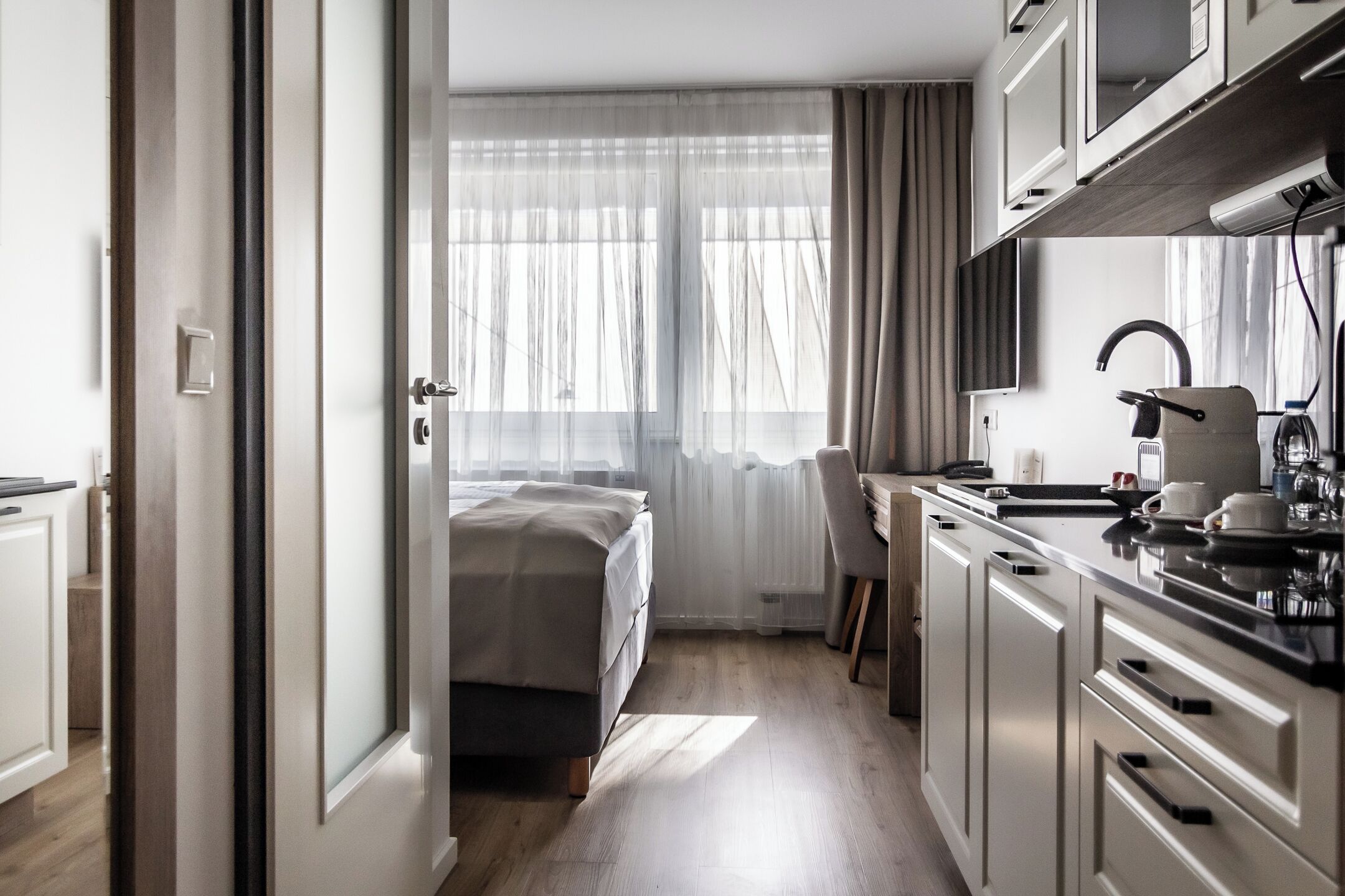 A modern hotel room with a bed, a desk, and a kitchenette.