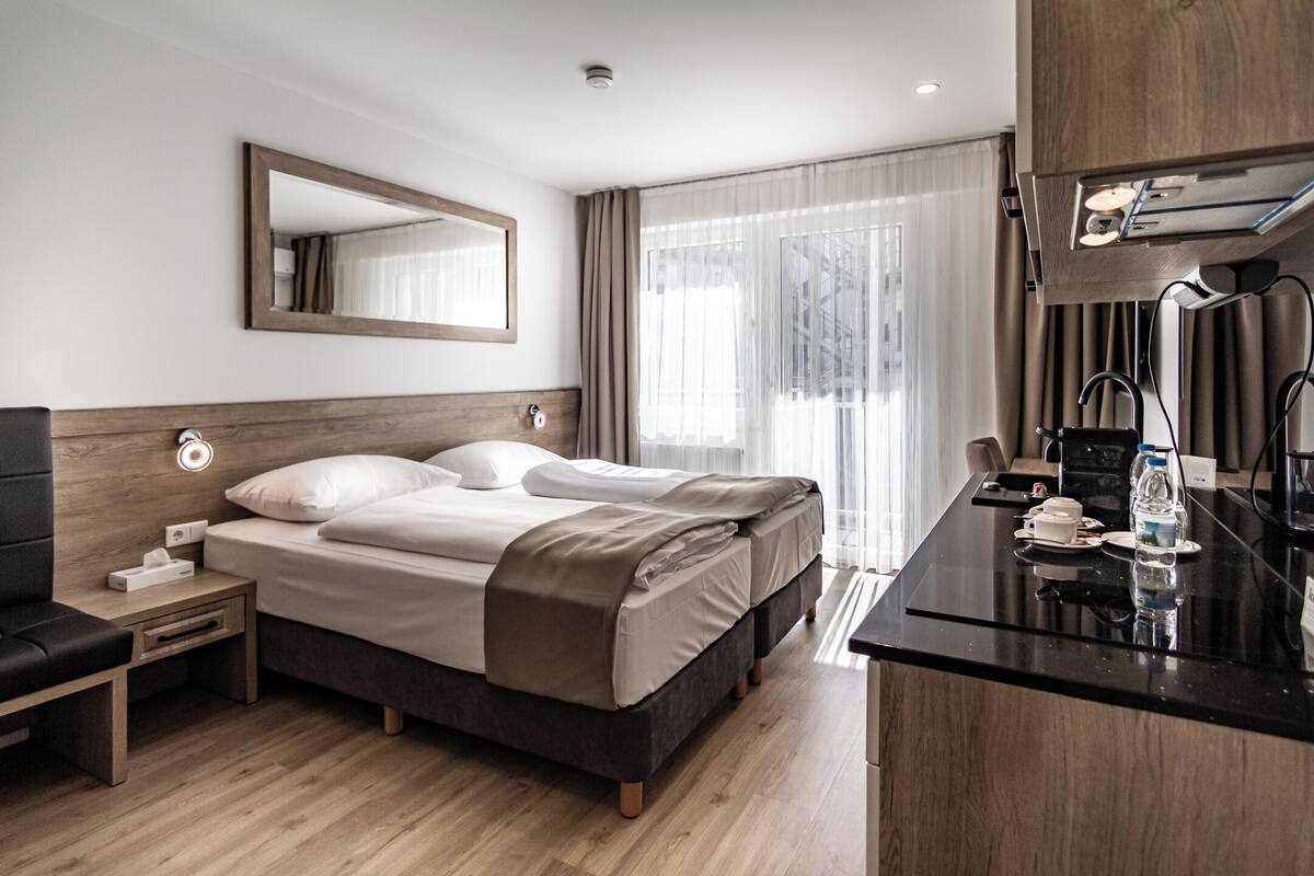 Une chambre d’hôtel moderne avec une tête de lit en bois, un grand lit, des tables de chevet, un bureau avec une chaise et une grande fenêtre avec des rideaux.