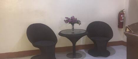 A room with two black armchairs, a small round table with a vase of flowers, a flat-screen TV mounted on the wall, and a fire extinguisher on the wall.
