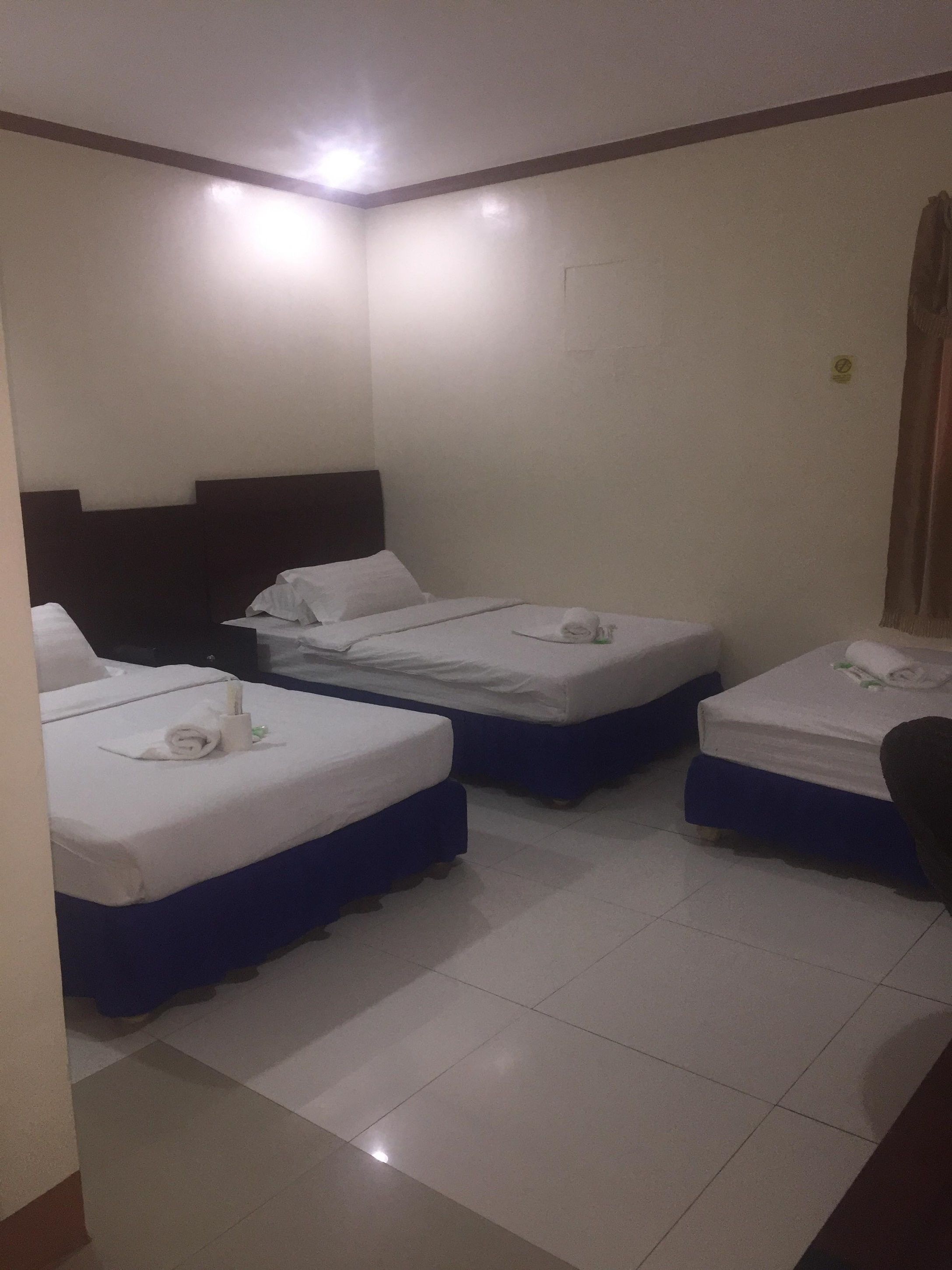 A hotel room with three beds arranged in a row, each with white bedding and blue headboards. The room has a tiled floor and a plain white wall with a small window.