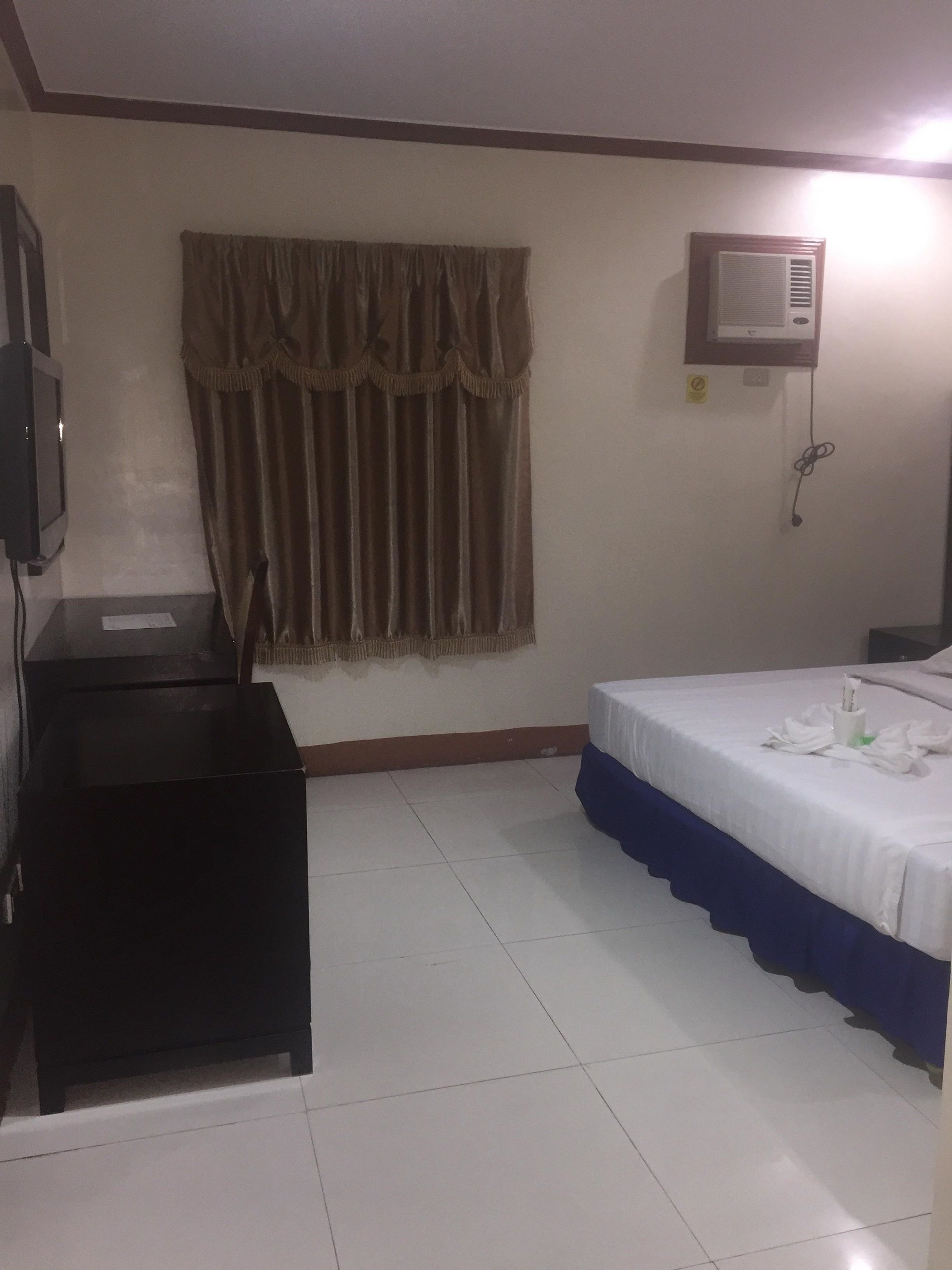 Quarto de hotel com cama, escrivaninha, televisão e janela com cortinas.
