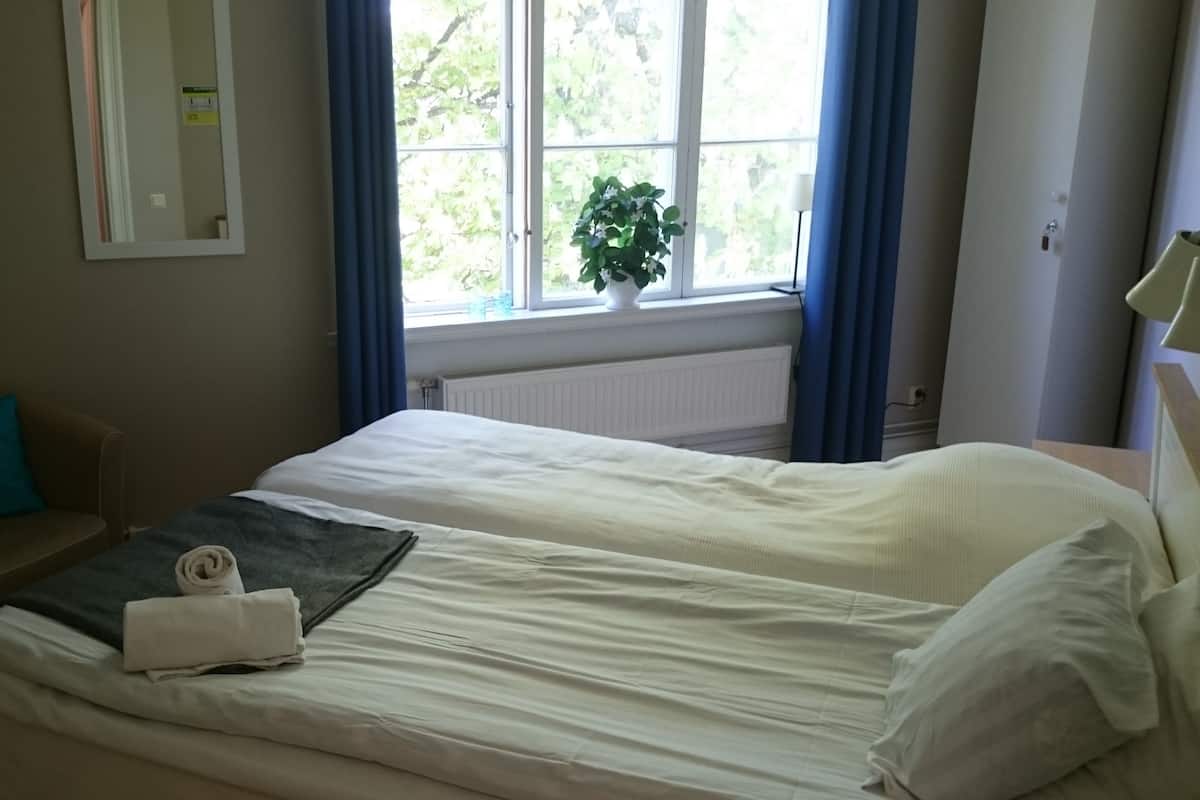 Une chambre d’hôtel avec un lit, une chaise, une fenêtre avec des rideaux, une plante et un radiateur.