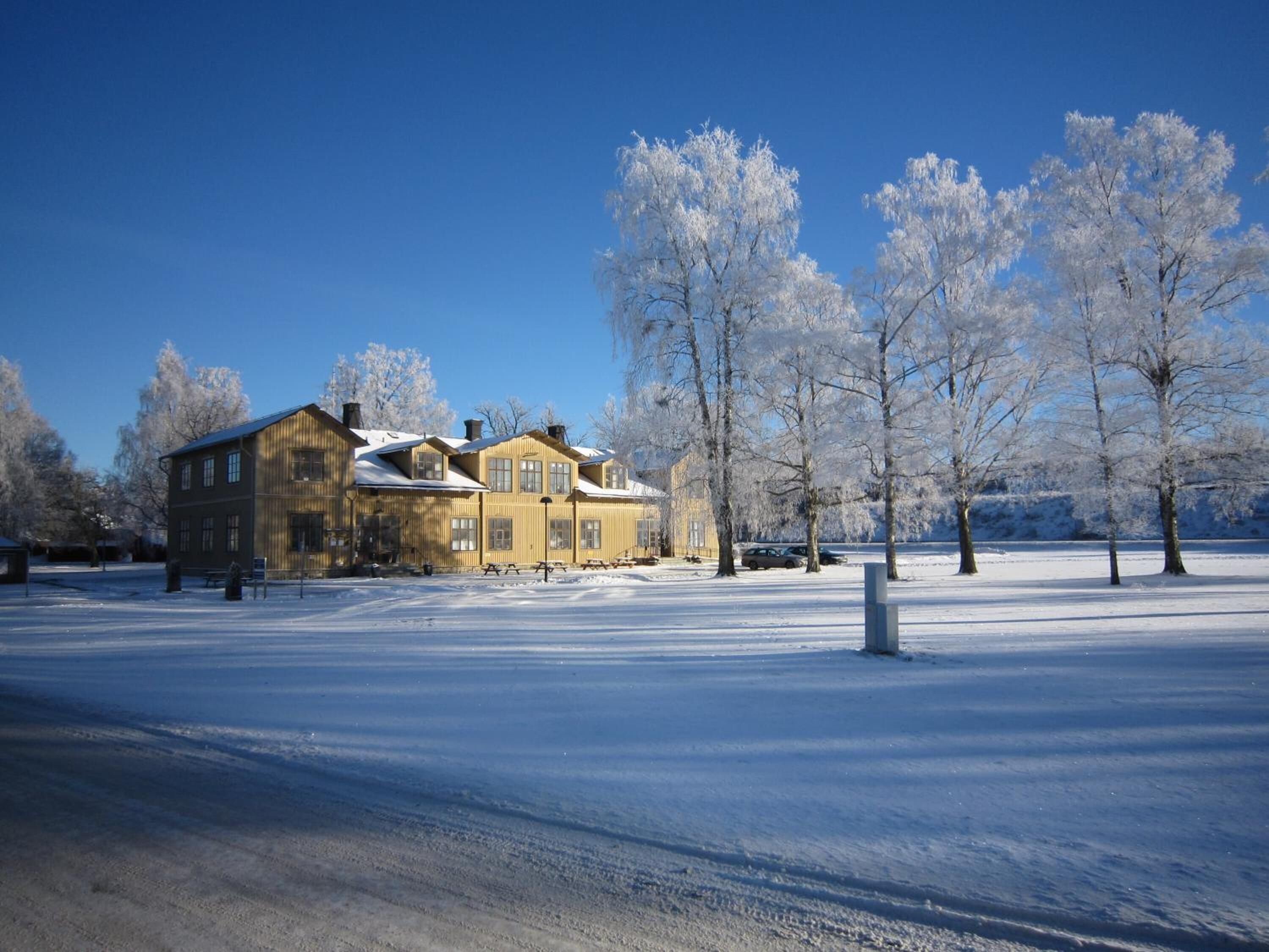 Ett stort, tvåvåningshus byggt av trä, med ett snötäckt tak, omgivet av ett snötäckt landskap och träd.