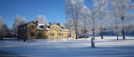 A large, two-story wooden house with a snow-covered roof, surrounded by a snow-covered landscape and trees.
