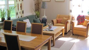 Een woonkamer met een bank, twee fauteuils, een salontafel en een eettafel. Er staan potplanten, een lamp en er hangen afbeeldingen in lijsten aan de muren.