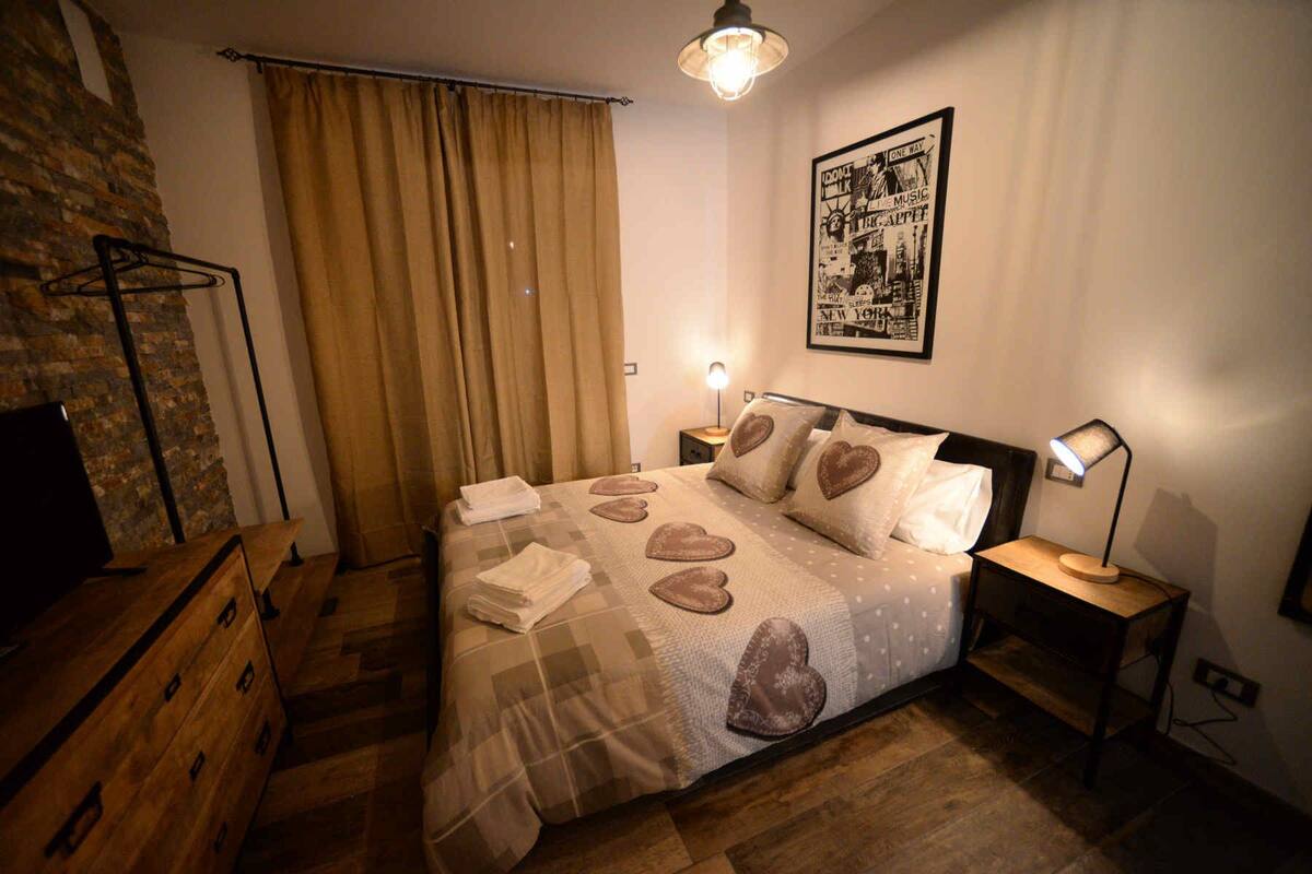 A bedroom with a bed, two bedside tables, a wall-mounted light, a framed poster, and a wooden dresser.