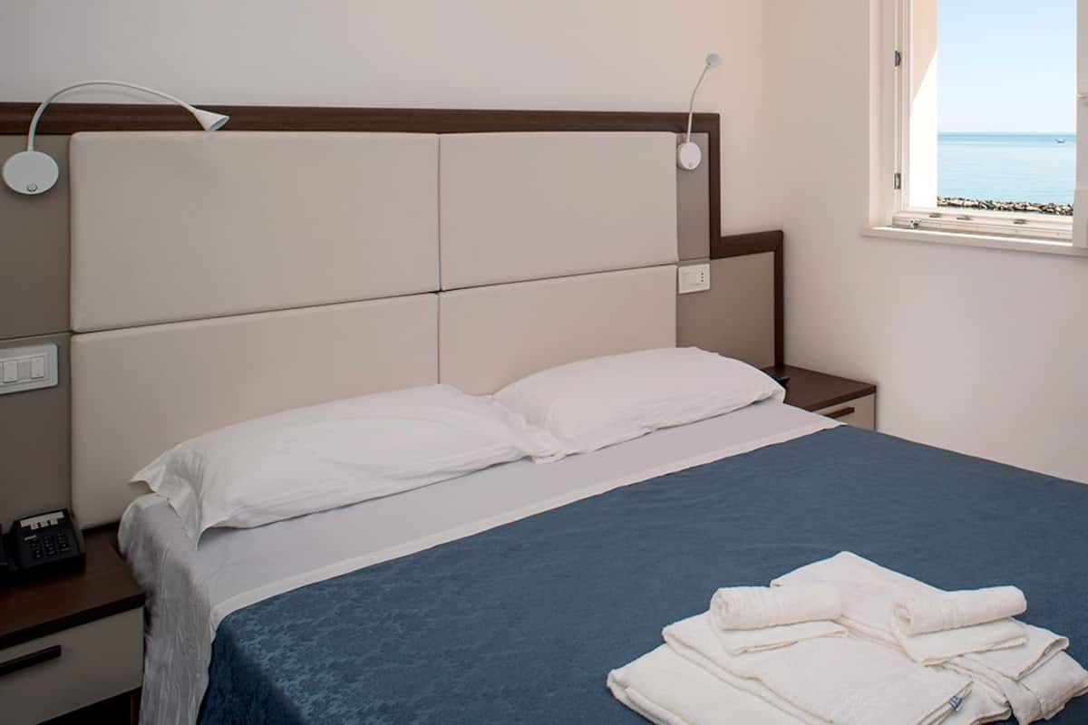 Un lit bien fait, avec du linge de lit blanc et une couvre-lit bleue, une table de chevet avec un téléphone, et une fenêtre donnant sur la mer.