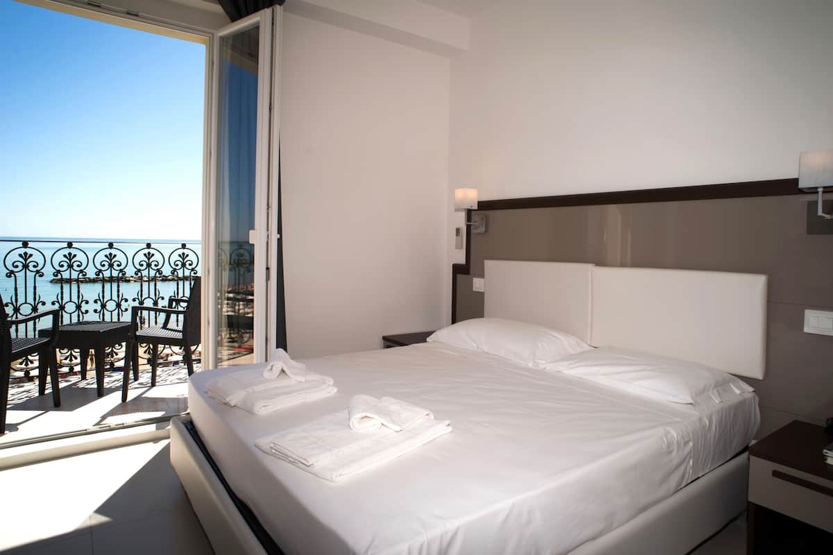 Une chambre d’hôtel avec un lit, une table de chevet, une fenêtre donnant sur la mer et un balcon avec des chaises.