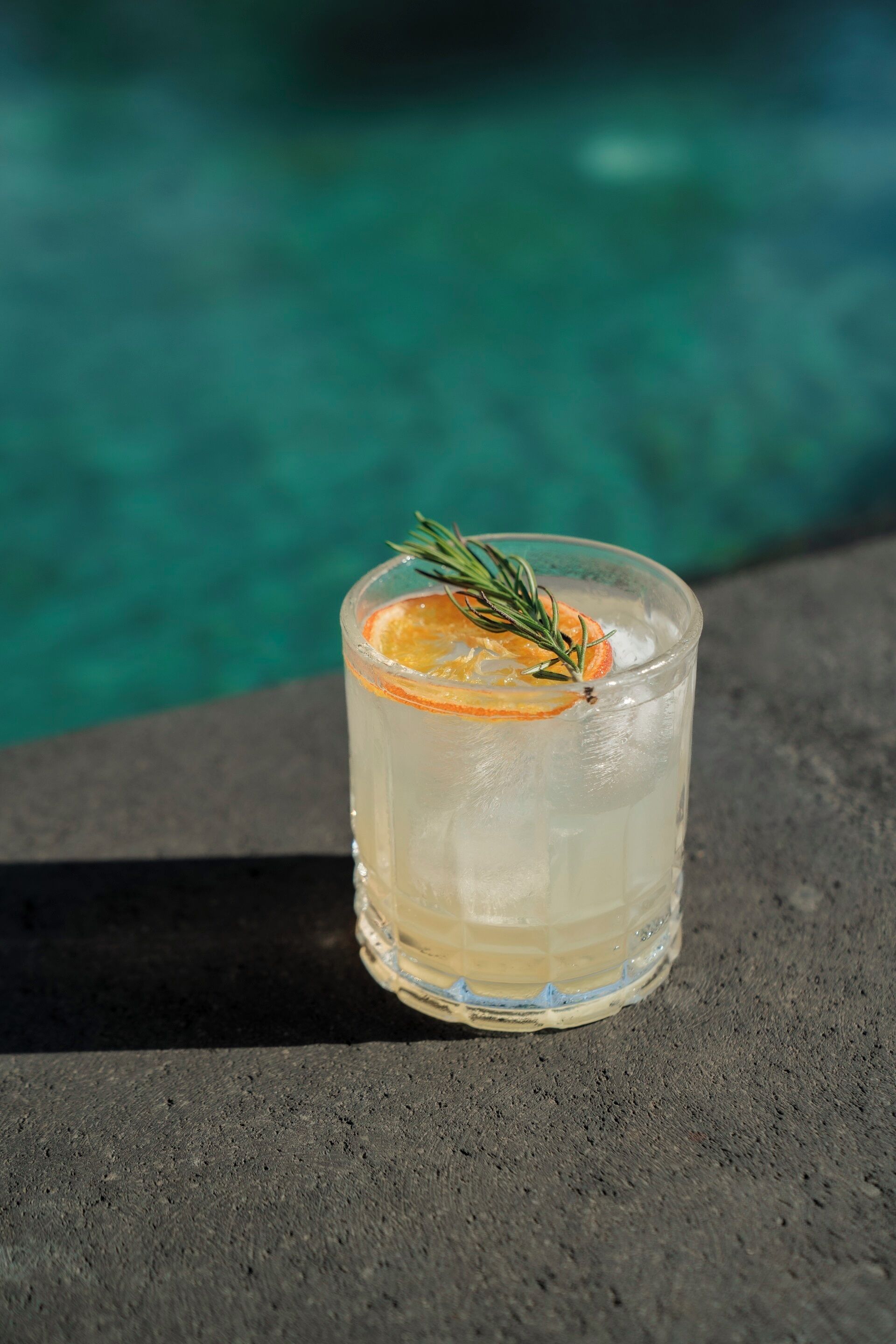 Un verre contenant une boisson claire, agrémenté d’une tranche d’orange et d’une branche de romarin, posé sur une surface sombre, avec une piscine en arrière-plan.