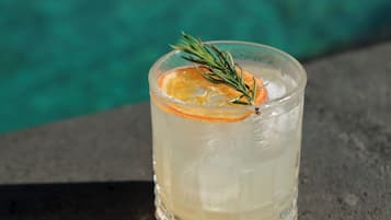 Un verre contenant une boisson claire, agrémenté d’une tranche d’orange et d’une branche de romarin, posé sur une surface sombre, avec une piscine en arrière-plan.