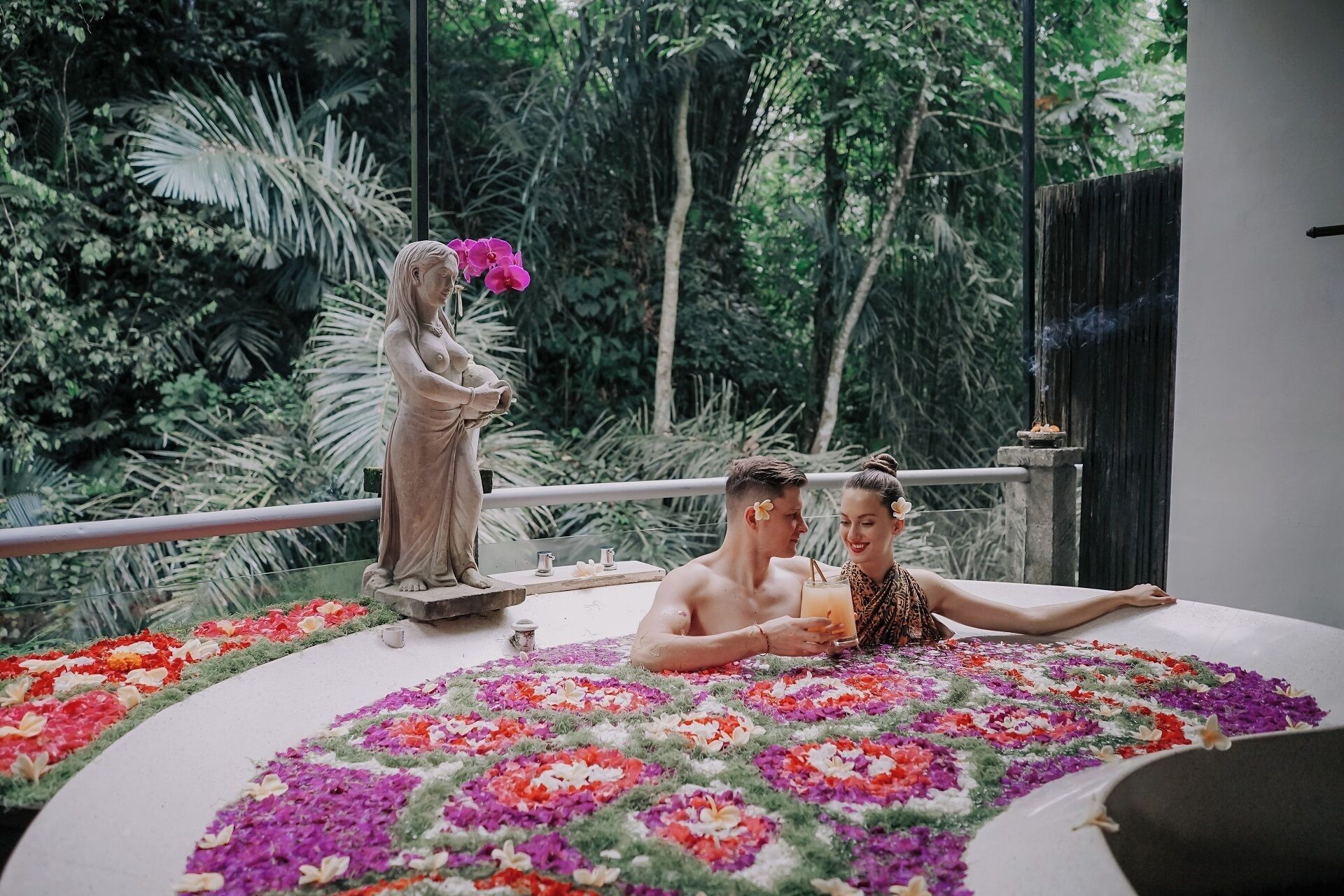 A tropical setting with a statue, a man and a woman on a bed, and a lush green background.