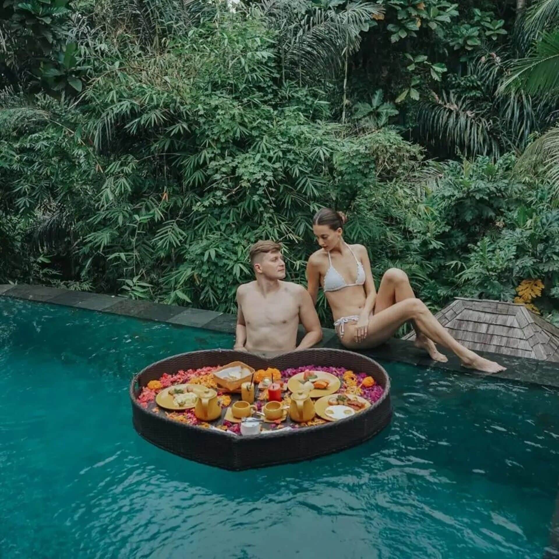 Two people sitting by a pool with a floating tray of food and drinks.