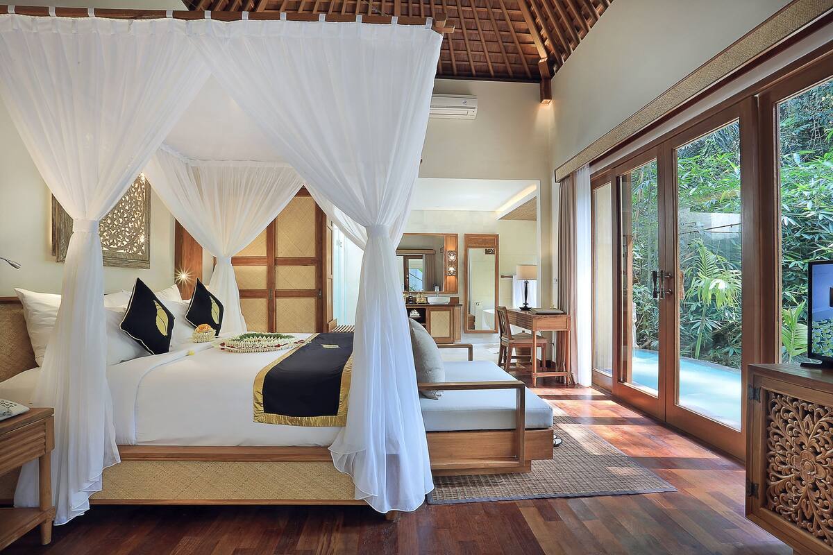 Une chambre à coucher avec un lit à baldaquin, un coin salon et de grandes fenêtres donnant sur l’extérieur.