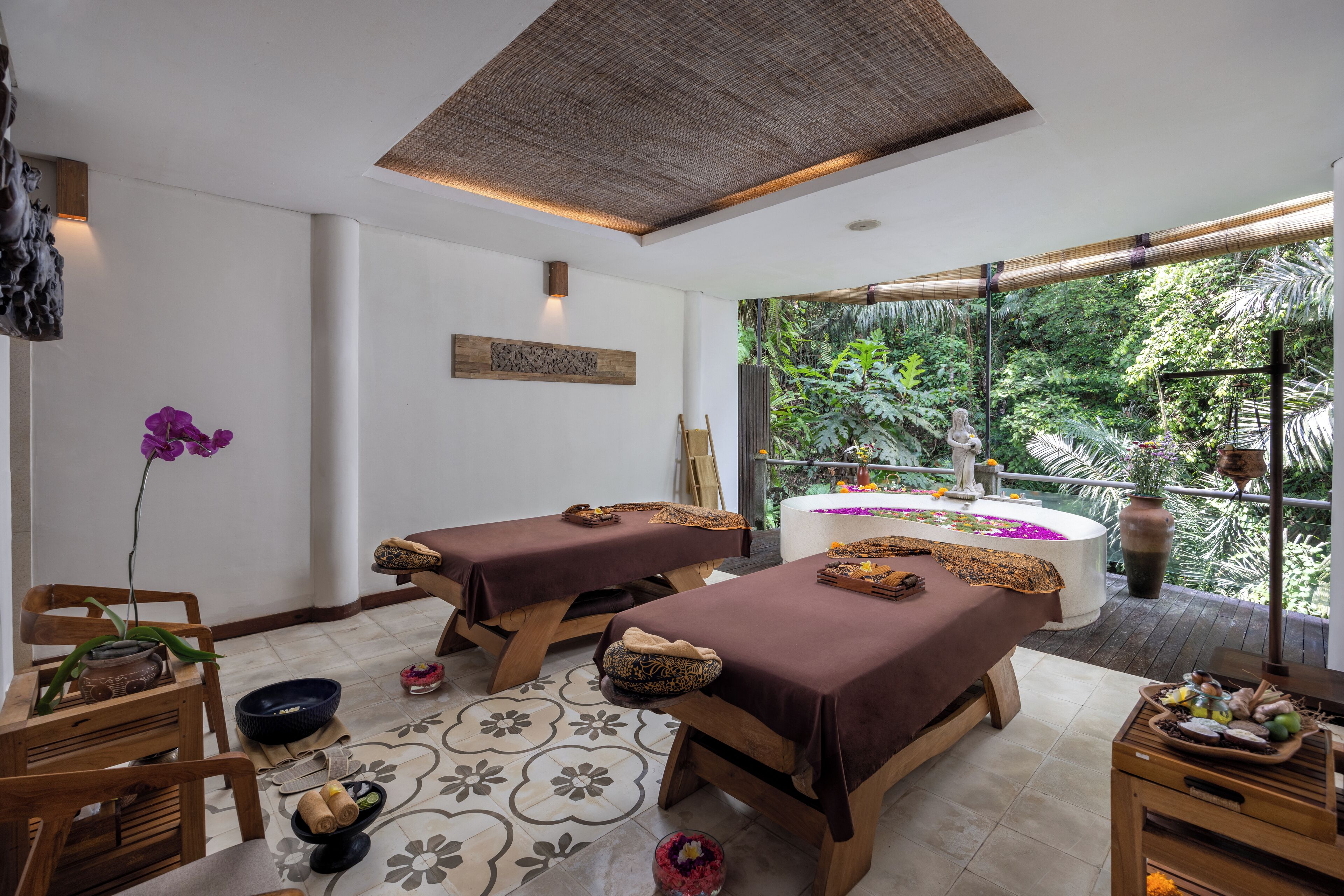 A spa room with two massage tables, a large window with a view of greenery, a decorative wall piece, and a small altar with flowers and candles.