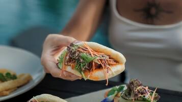 A person holding a sandwich with various toppings, a plate with another sandwich, and a glass of beverage on a table.