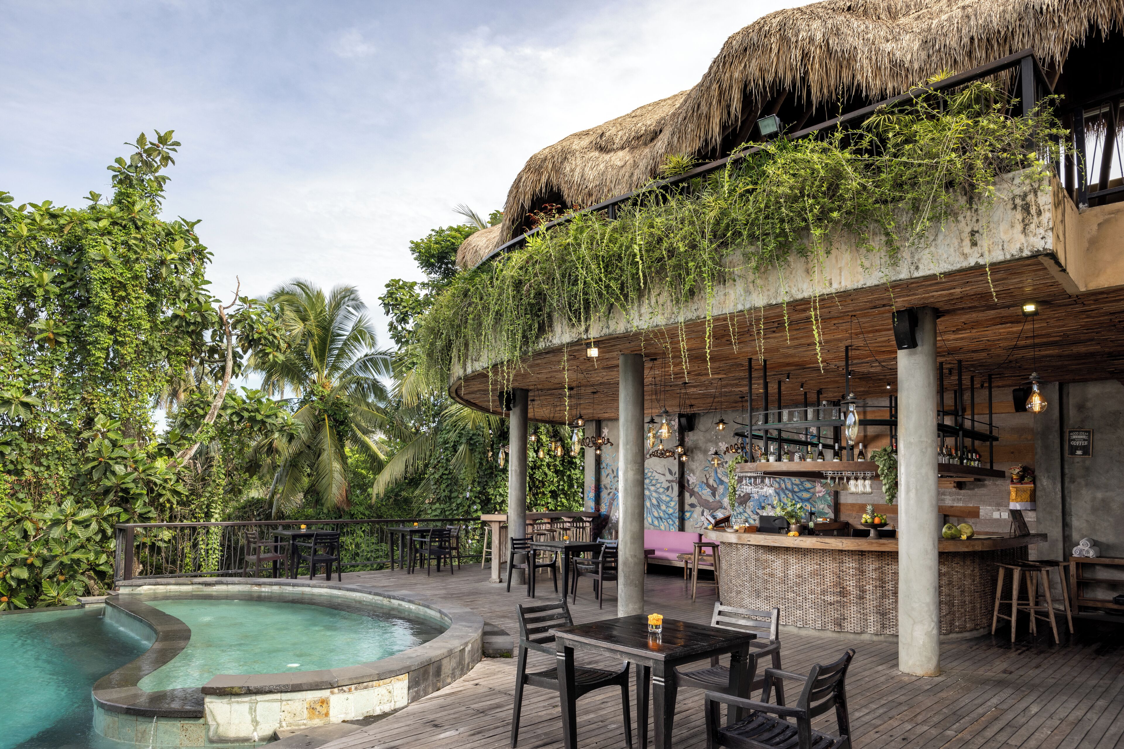 Un área junto a la piscina con bar y asientos, rodeada de vegetación exuberante y una estructura con techo de paja.